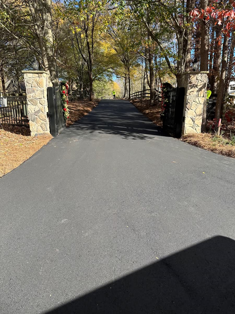 Long asphalt driveway curving between tall evergreen trees and stone pillars